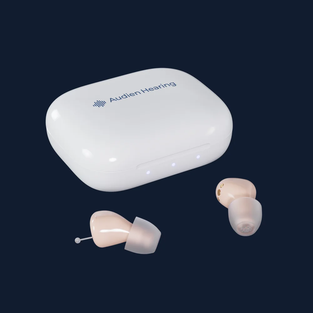 An Audien Atom Air charging case with the pair of beige in-ear hearing aids beside it, photographed on black