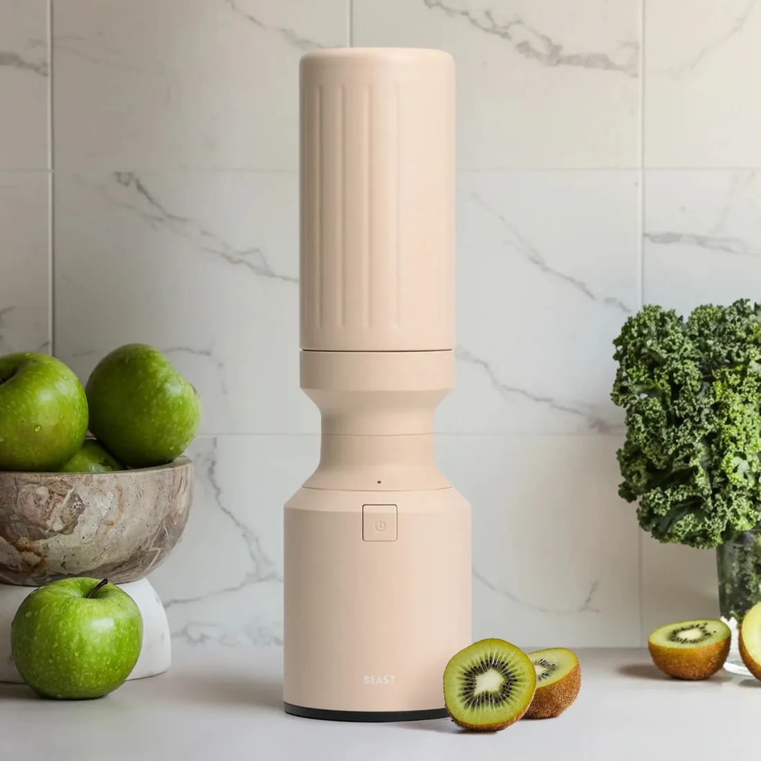 A sand coloured Beast blender on a kitchen counter next to a bowl of apples, kiwi and kale