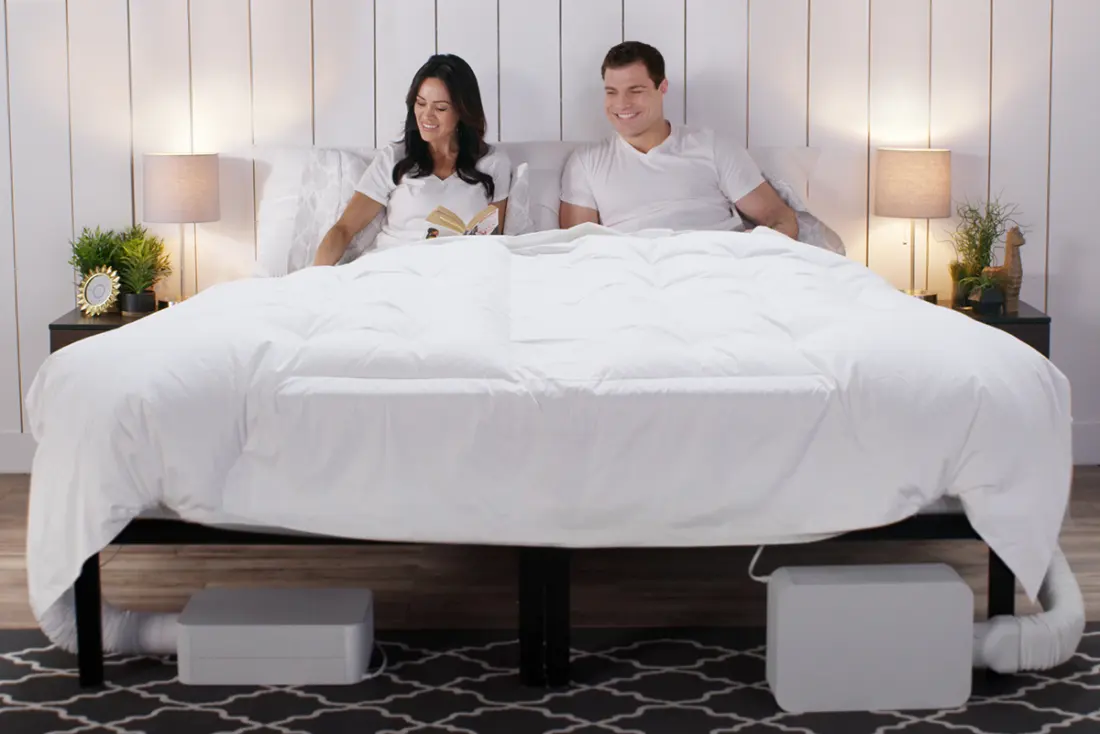 A couple sitting up in bed reading, with two BedJet units visible on the floor at the foot of the bed.