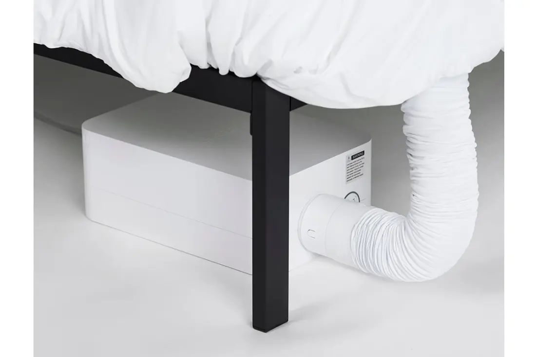 A BedJet unit on the floor beside a bed frame, with its hose running up into the bedding.