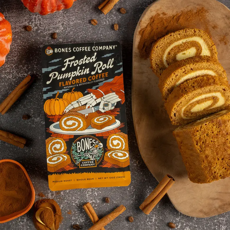 A bag of Bones Coffee Frosted Pumpkin Roll flavored coffee laid on a slate surface beside three slices of pumpkin roll on a wooden board, with cinnamon sticks, whole coffee beans and a bowl of ground spice