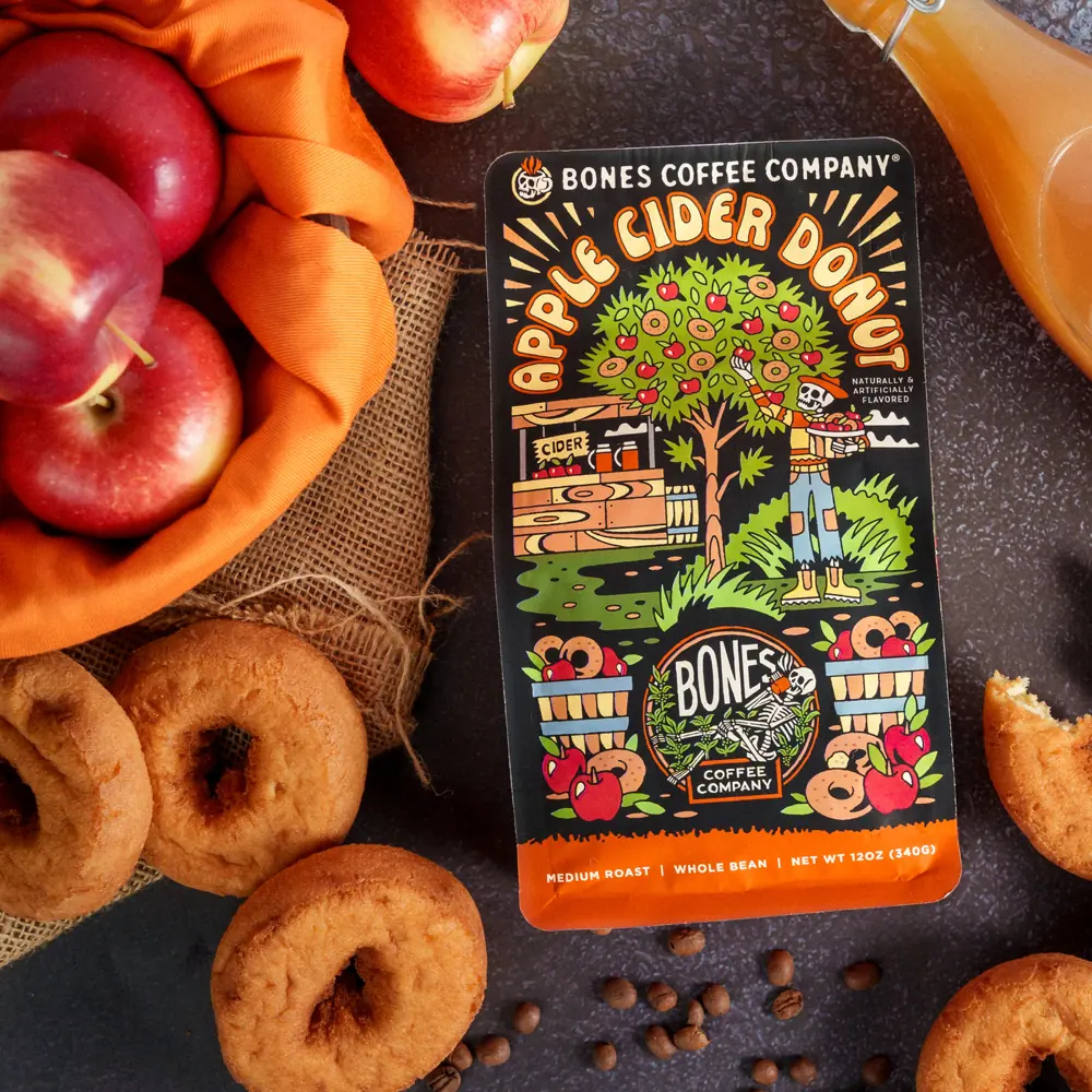 Apple Cider Donut coffee bag on a dark surface surrounded by cider donuts, red apples and a jug of cider