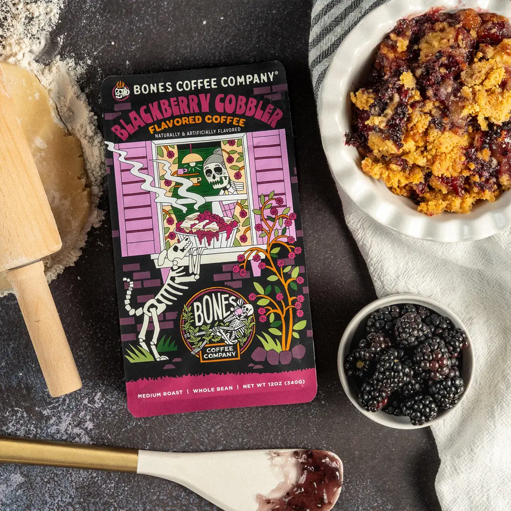 Blackberry Cobbler coffee bag on a dark surface beside a dish of blackberry crumble, fresh blackberries and a rolling pin