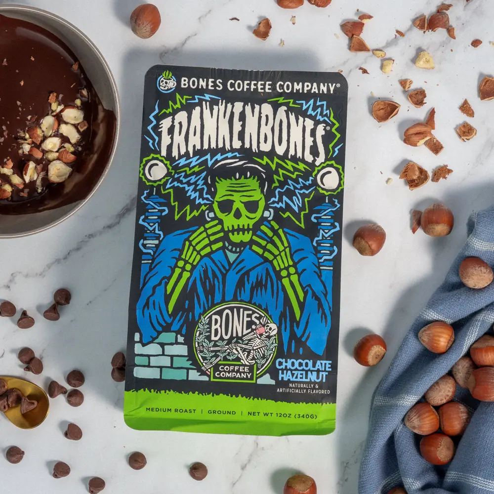 Frankenbones chocolate hazelnut coffee bag on a marble surface with a bowl of melted chocolate and scattered hazelnuts