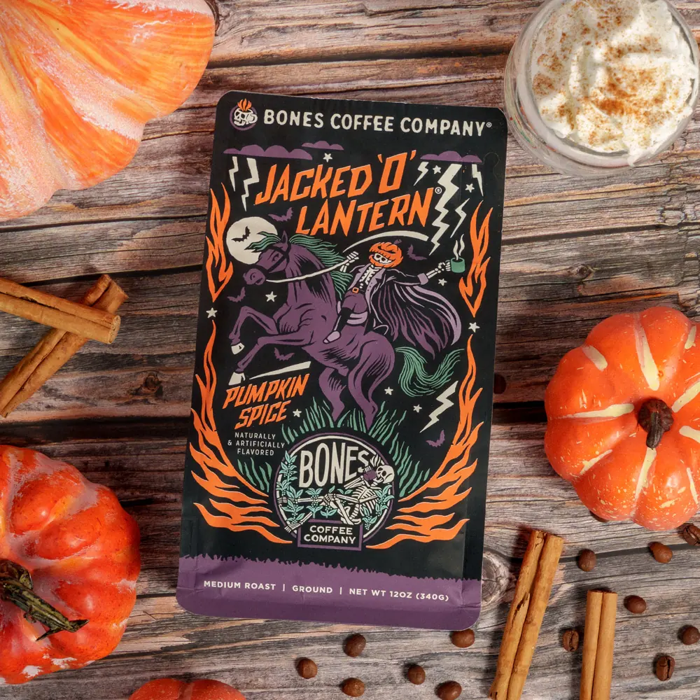 Jacked O Lantern pumpkin spice coffee bag on a wooden table with small pumpkins, cinnamon sticks and a topped latte