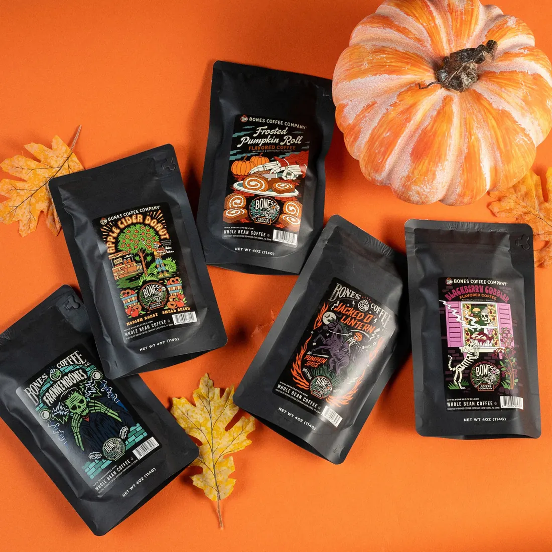 Five bags of Bones Coffee fall flavored coffee arranged on an orange background with a white pumpkin and autumn leaves