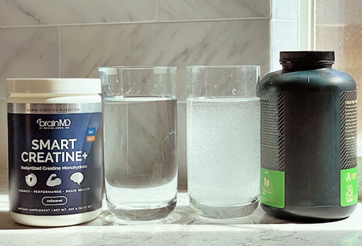 Two glasses of water side by side, the left one clear next to a tub of BrainMD Smart Creatine, the right one cloudy next to an unbranded black tub of conventional creatine