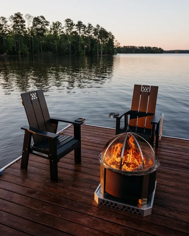 A Breeo X19 fire pit burning on a wooden dock at dusk with two chairs beside it