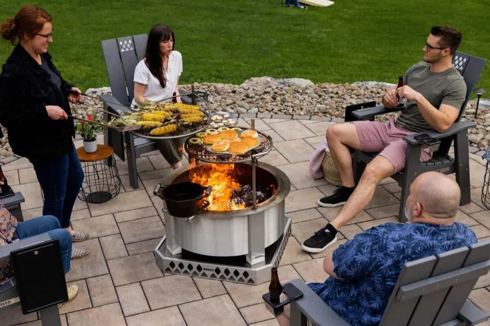 A group of friends gathered around a lit Breeo X30 fire pit with grilling attachments in place