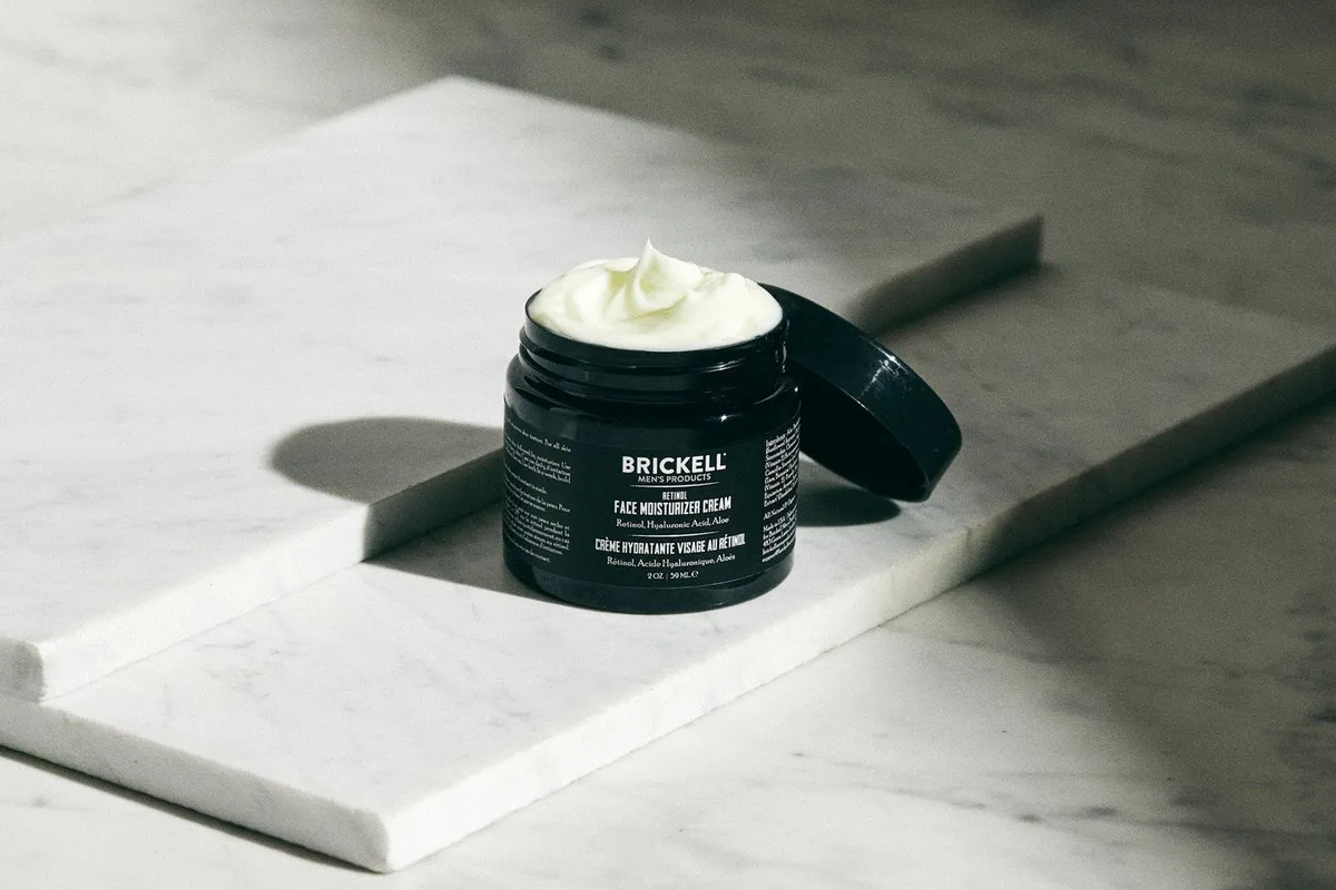 An open Brickell face moisturizer cream jar on a marble slab, lid resting beside it