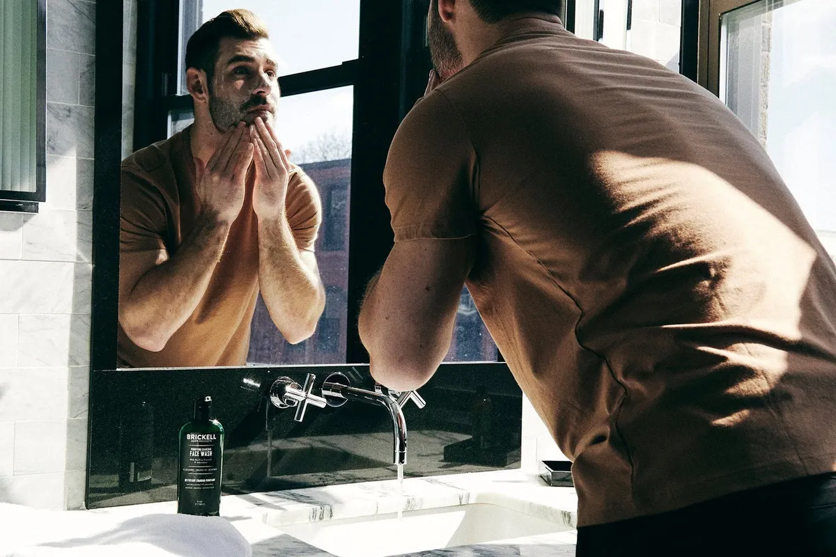 A man washing his face at a bathroom sink, seen from behind with his reflection in the mirror