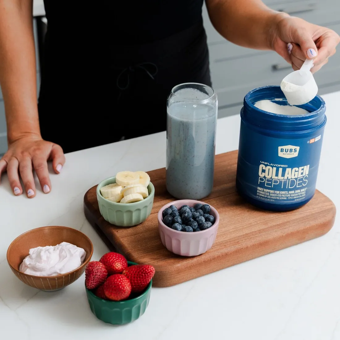Hands scooping collagen powder into a blender jar beside strawberries, blueberries and banana on a wooden board.