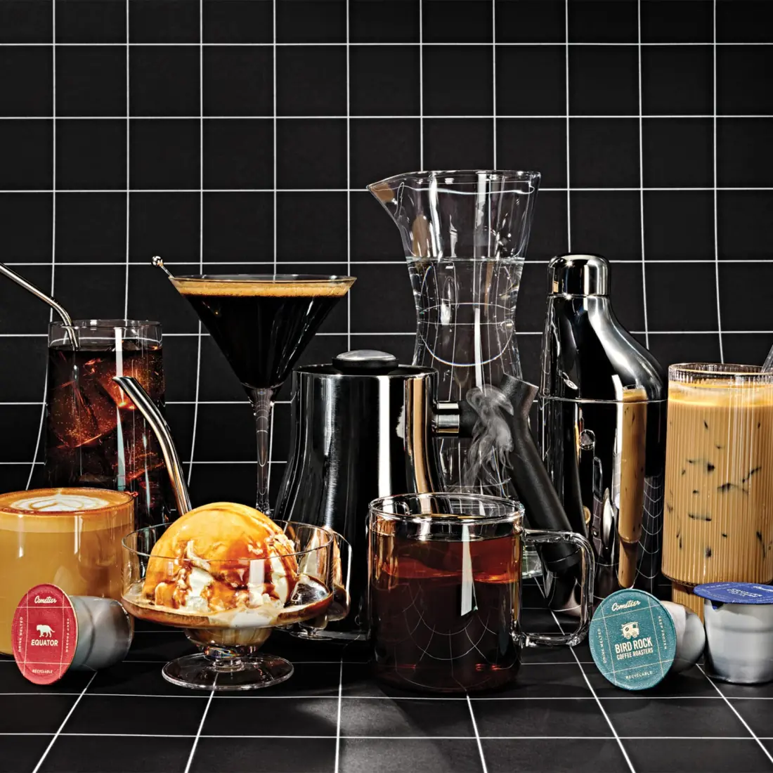 Iced coffees, an espresso martini, a hot mug, a latte and an affogato arranged on a black tiled surface with Cometeer capsules in front