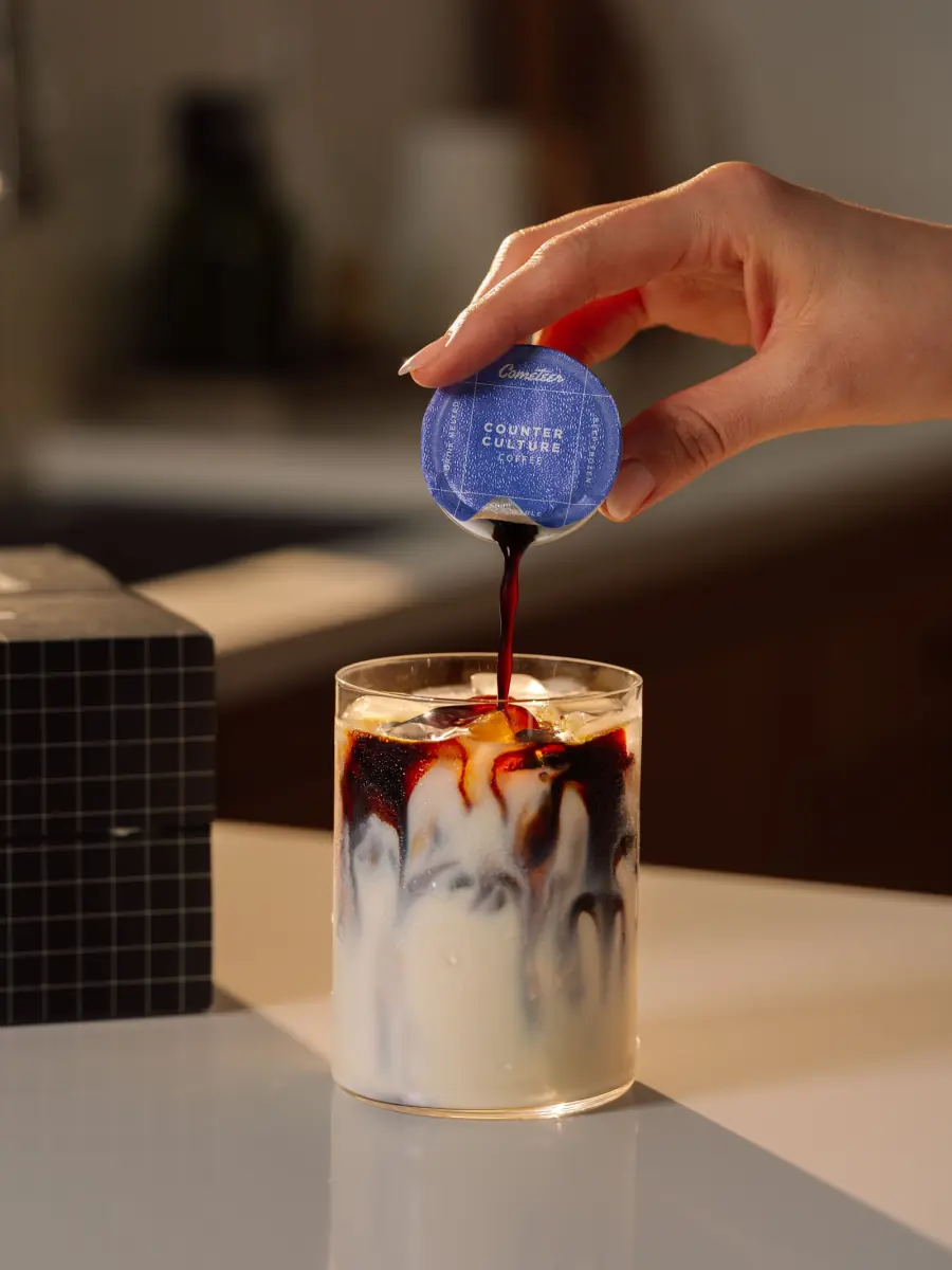 A thawed Cometeer capsule being poured into a glass of milk over ice, the coffee falling through the milk in dark ribbons