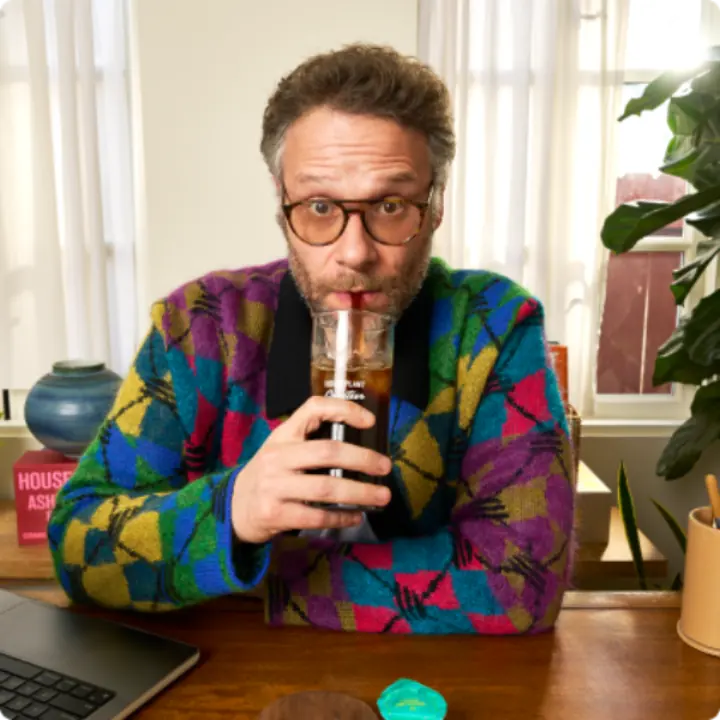 Seth Rogen drinking an iced coffee through a straw at a desk, with a Cometeer capsule in front of him