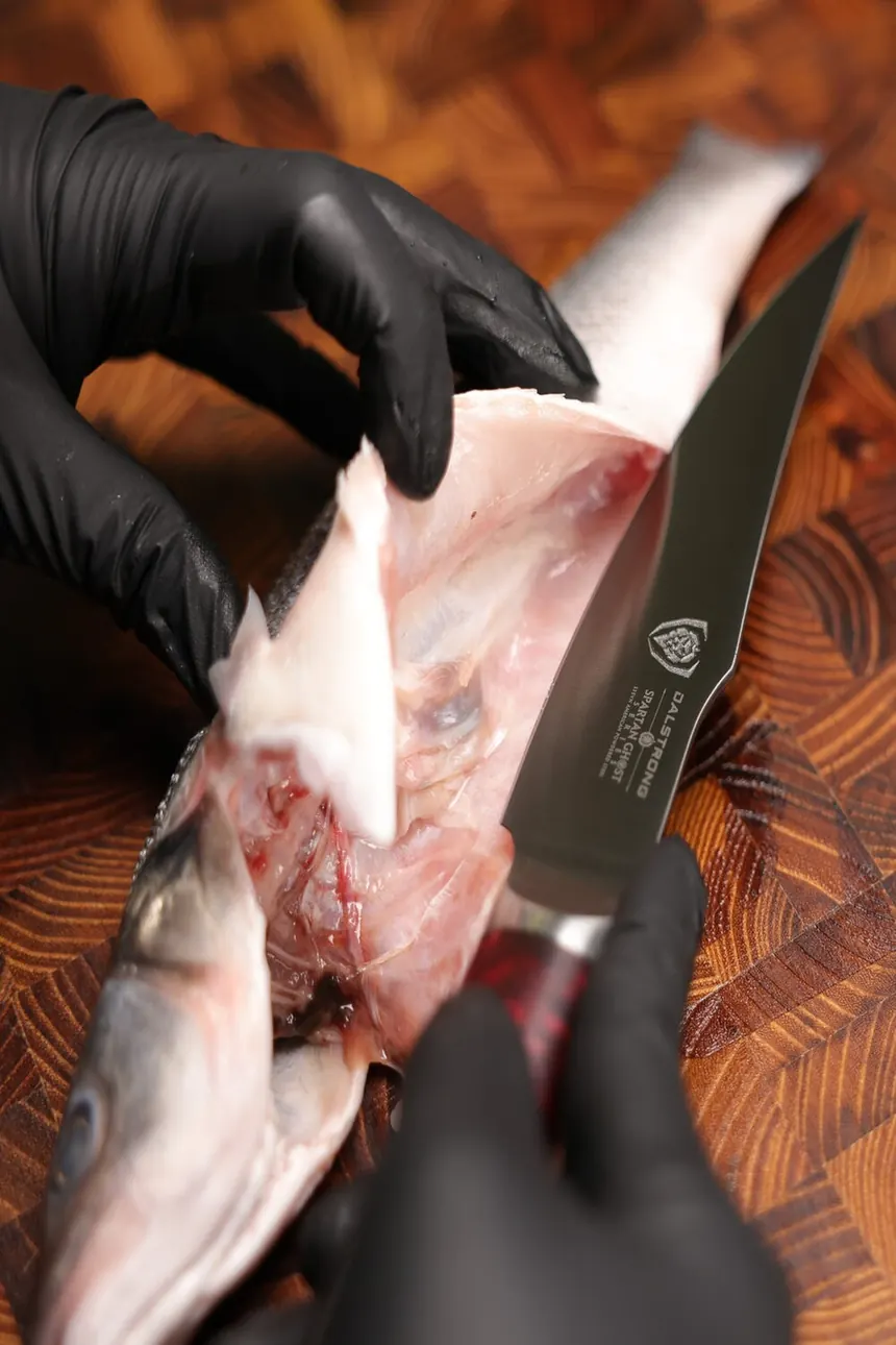 A gloved hand filleting a fish on a wooden board with the Spartan Ghost curved fillet knife