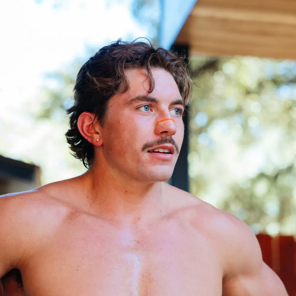 A man outdoors in bright sun wearing an orange nasal strip, skin visibly wet with sweat
