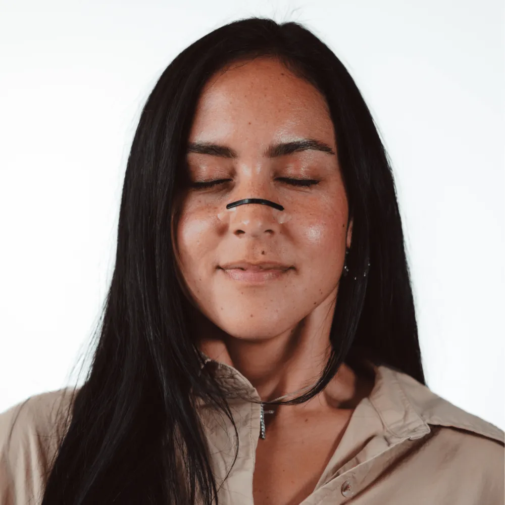 A woman with her eyes closed wearing a black nasal strip, photographed against a white background