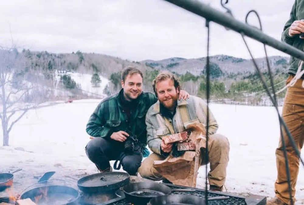 Brothers Stephen and Chris Muscarella outdoors in winter, crouched by a fire with cast iron cookware