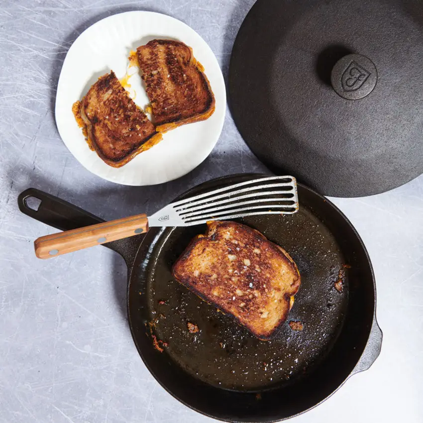 A grilled cheese sandwich lifting cleanly out of a dark seasoned cast iron pan, with a second sandwich plated beside it