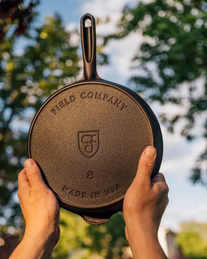 A person holding up a Field cast iron skillet outdoors, underside forward, showing the maker's mark and the thin rim
