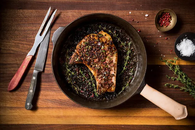 A seared pork chop resting in a dark seasoned cast iron pan on a wooden board with a carving knife