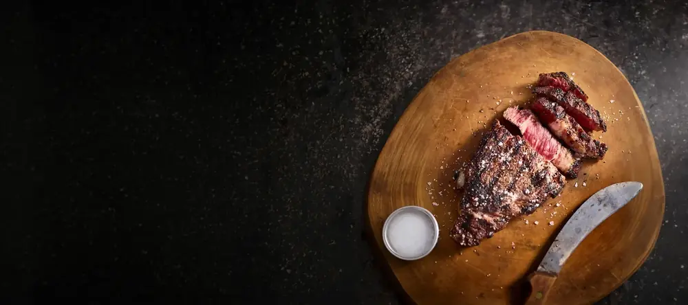 A cooked steak resting on a wooden board with a knife and coarse salt