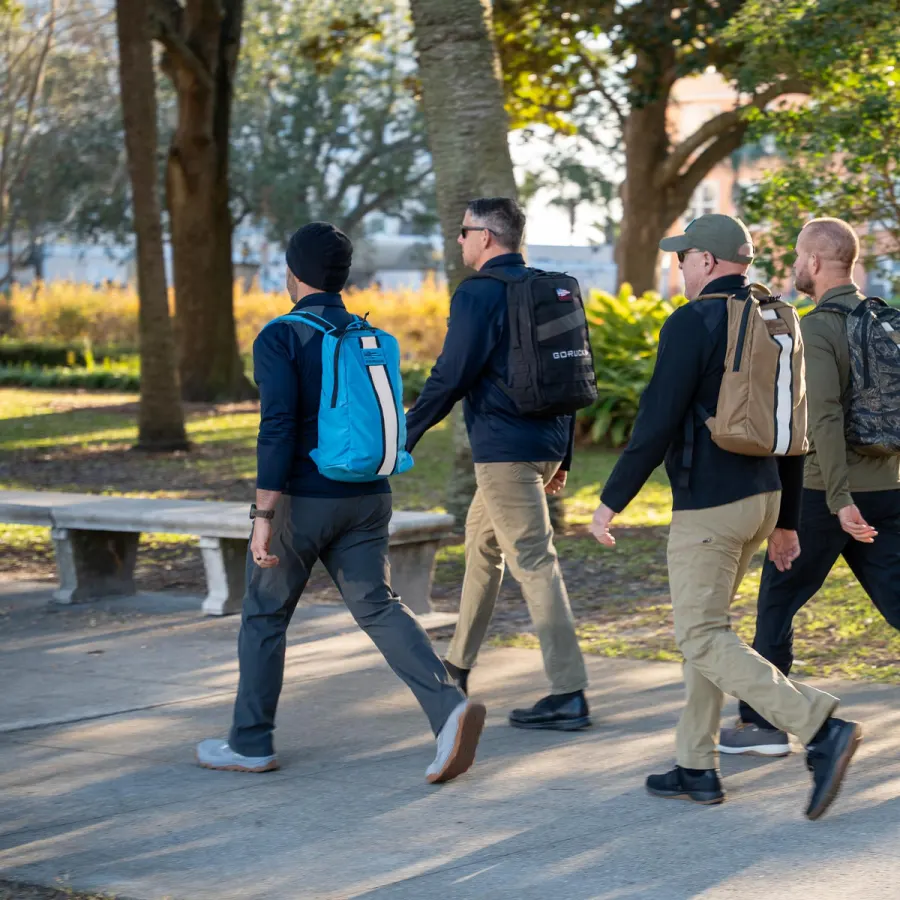 A group of people in ordinary clothes walking through a park wearing loaded rucksacks