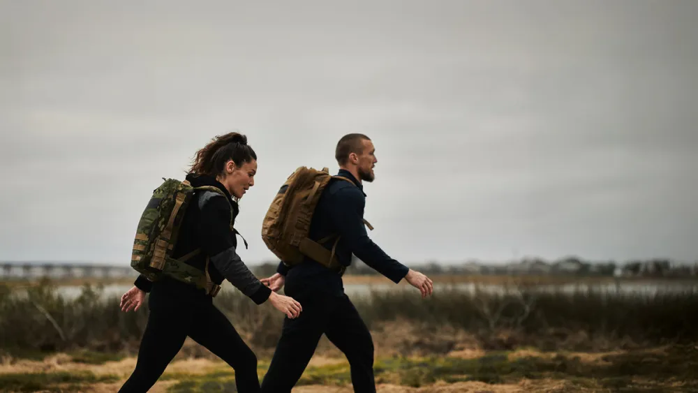 Two people walking in profile with loaded rucksacks, shoulders back and heads stacked over their shoulders