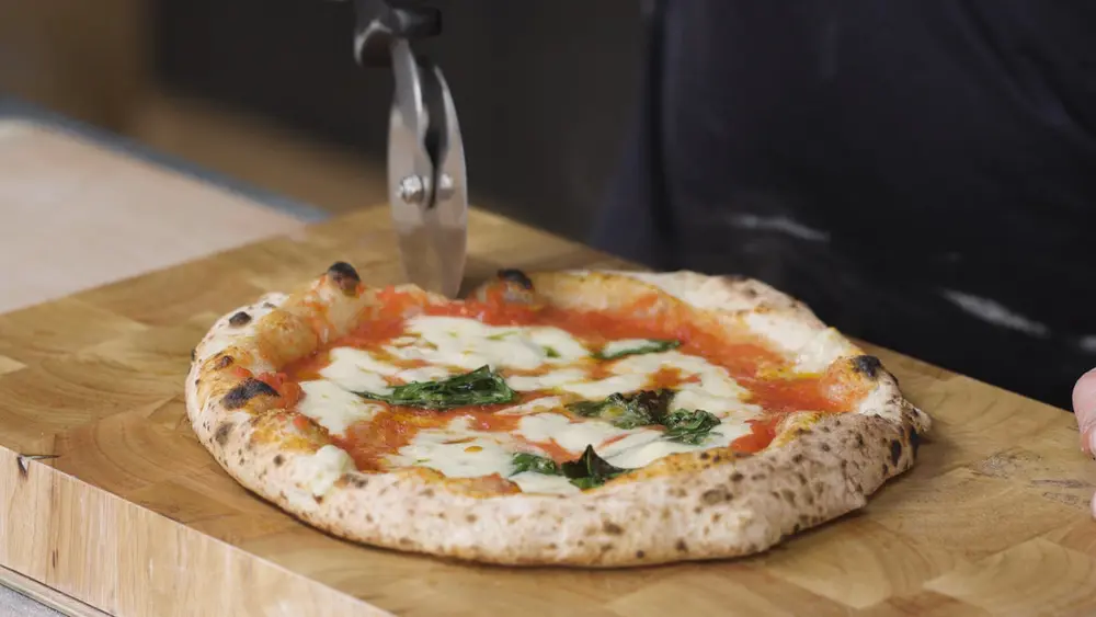 A finished Neapolitan pizza with a puffed, spotted crust being cut on a wooden board.