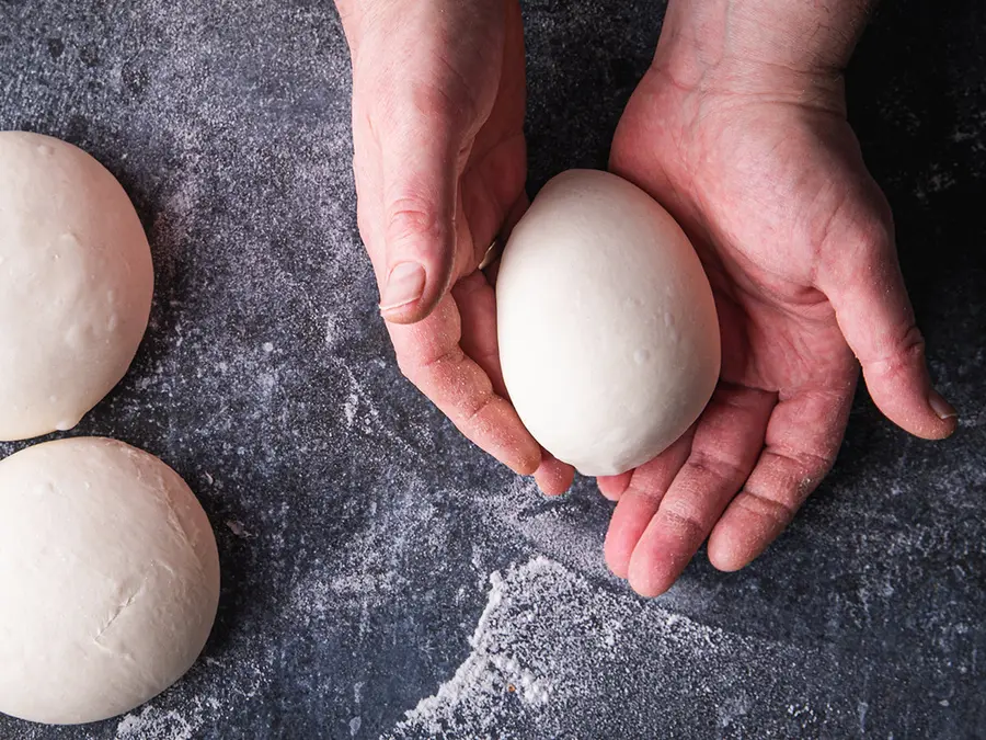 Two hands cradling a proofed pizza dough ball above a floured worktop.