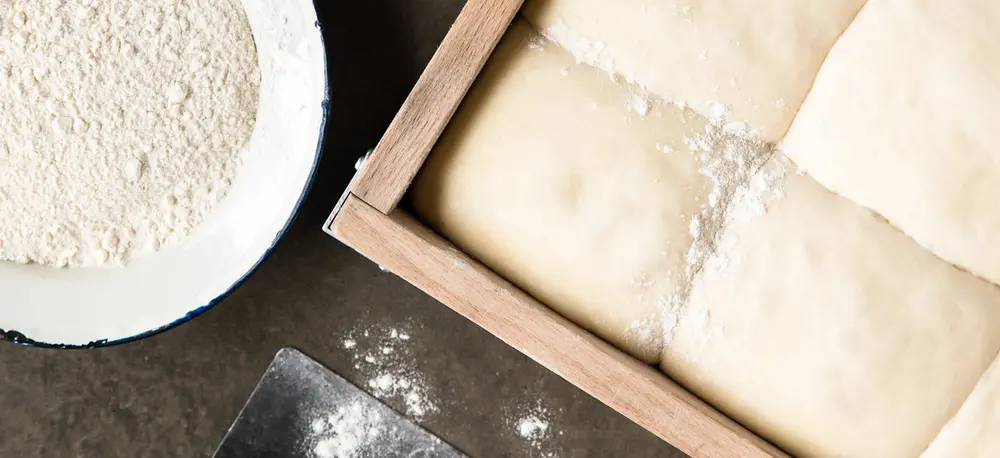 A dough tray of proofed pizza dough balls beside a bowl of flour and a dough cutter.