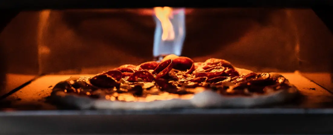 A pizza baking on the stone floor of a Gozney oven with the flame rolling across the chamber above it.