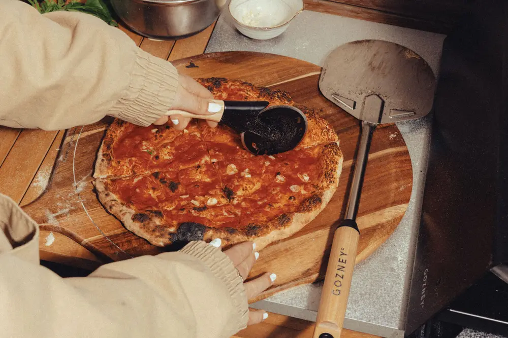 A stretched pizza base being dressed with sauce on a wooden board, next to a Gozney peel and oven.