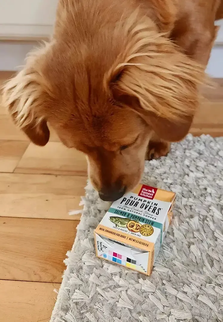 A golden retriever pressing its nose against a closed cardboard box of dog food topper on a rug