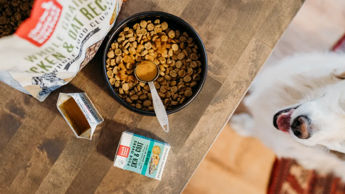 Overhead view of a bowl of kibble with a spoon of topper, boxes of pour overs and a bag of food on a wooden table, with a dog's nose entering the frame