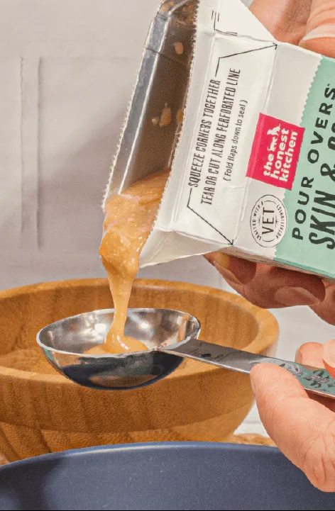 A hand tipping a carton of stew topper over a measuring spoon and a dog bowl