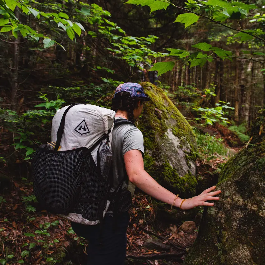 A hiker in dense green forest wearing a light pack, steadying herself on a mossy boulder.