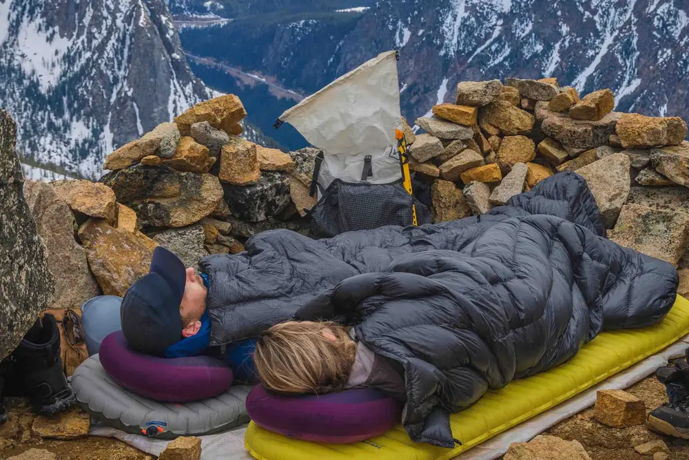 Two people lying in quilts on sleeping pads at a rocky high camp with mountains behind.