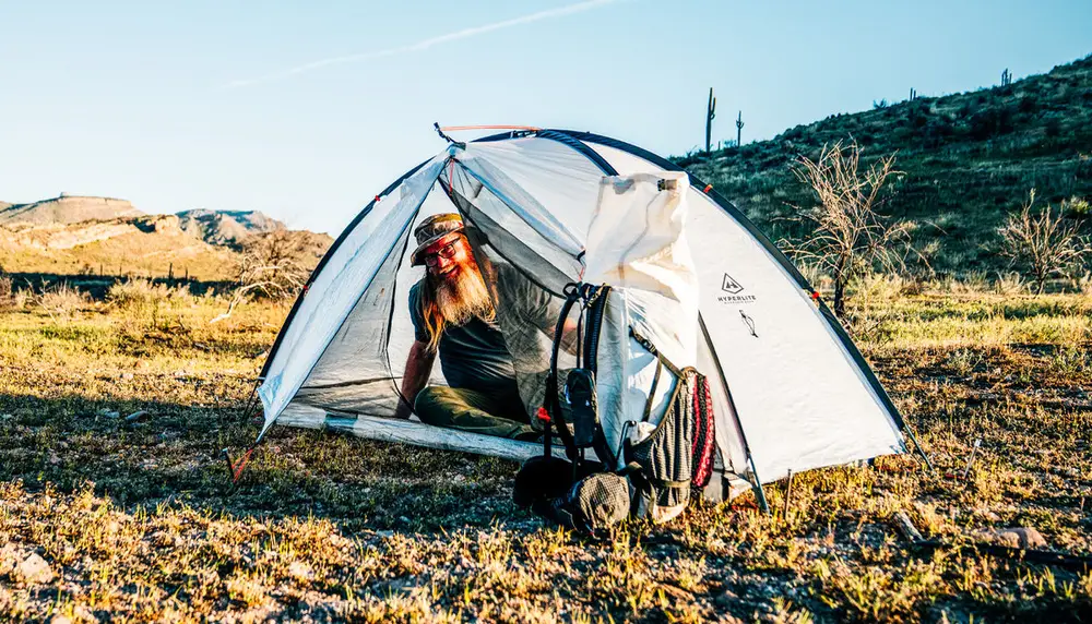 A camper sitting in the door of a white shelter at first light in dry mountain terrain.