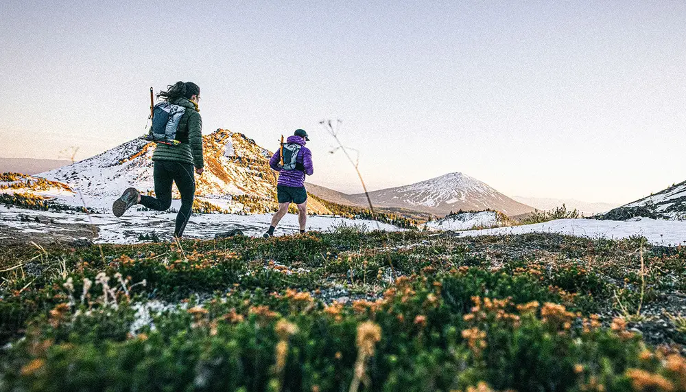 Two hikers with light packs crossing an alpine meadow with snow-covered peaks behind them.