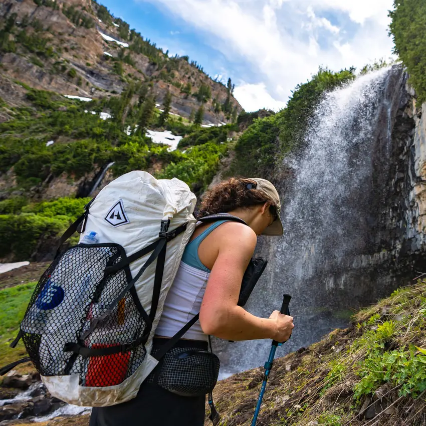 A hiker carrying a white Windrider pack with mesh pockets on a steep green slope beside a waterfall.