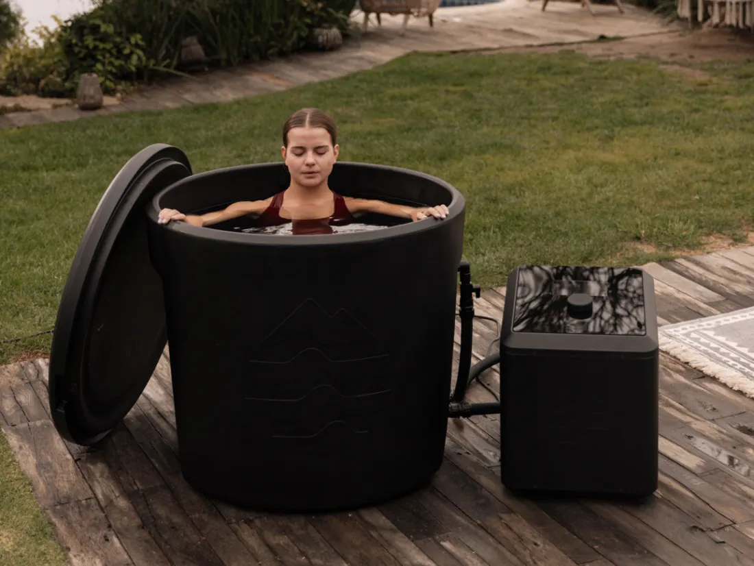 A woman sitting in an Ice Barrel on a wooden deck with the Chiller unit beside it