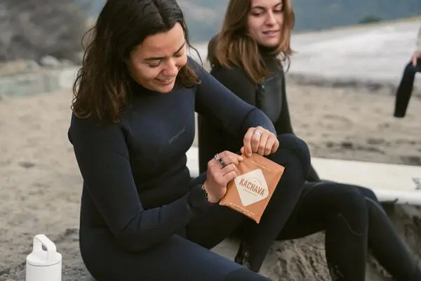 Two people in wetsuits on a beach opening a single serve sachet