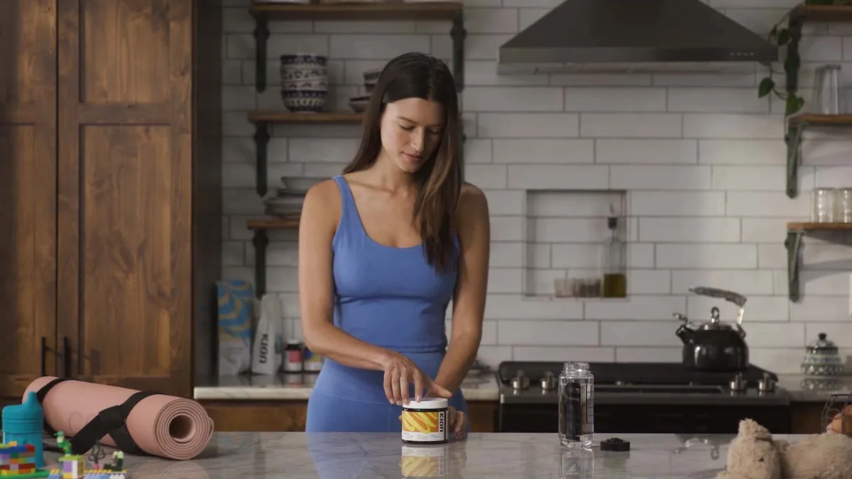 A woman scooping Kion Aminos powder into a shaker bottle in a kitchen, next to a rolled yoga mat