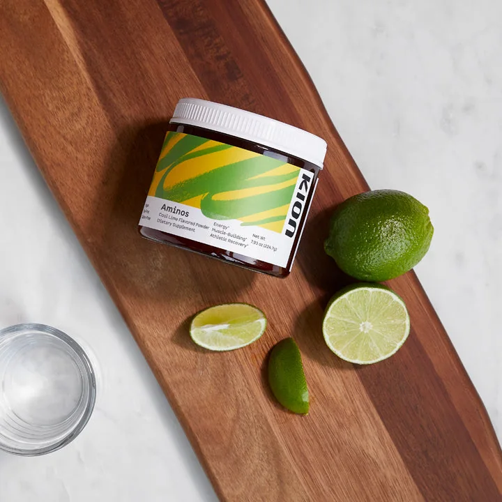 A tub of Kion Aminos Cool Lime powder on a wooden board with fresh limes and a glass of water