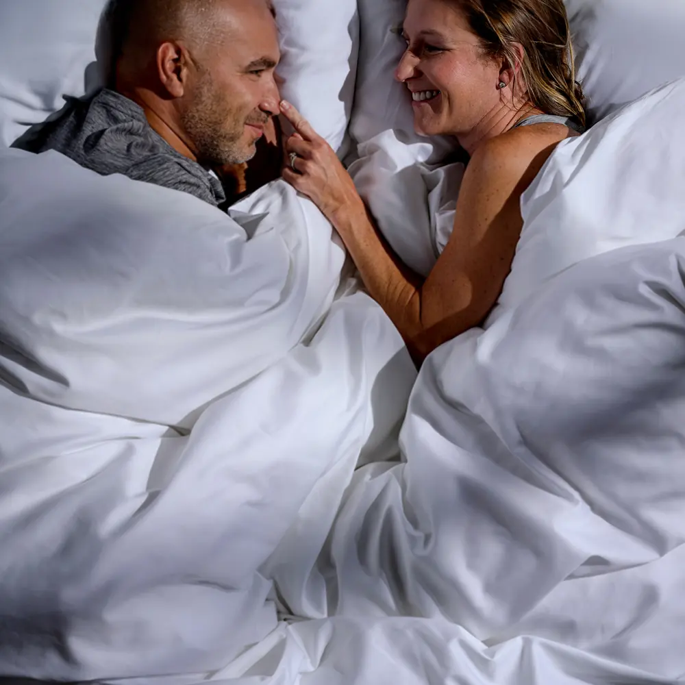 A couple lying face to face in bed under a white comforter, seen from above