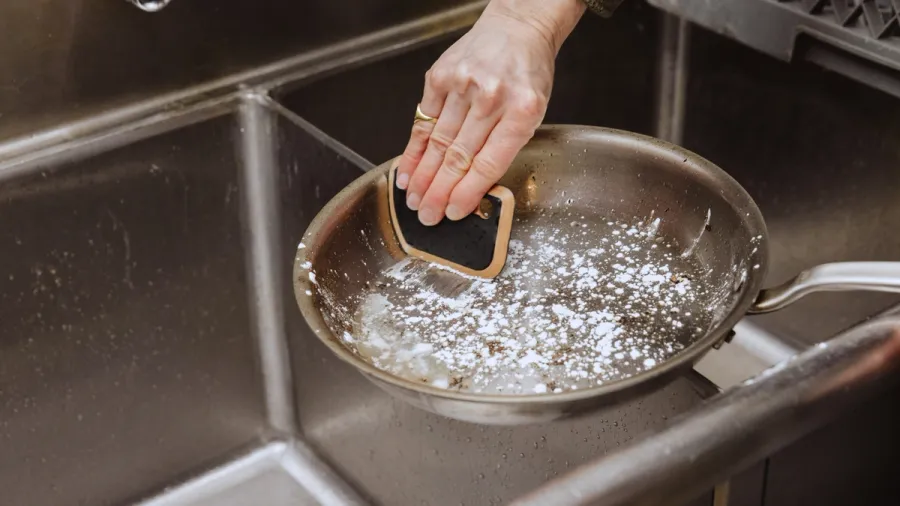 A hand cleaning a stainless steel frying pan with a sponge in a sink
