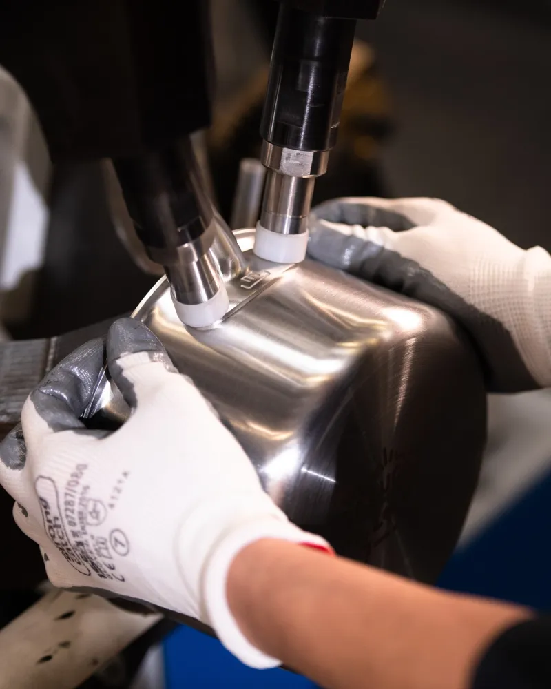 Gloved hands holding a stainless steel pan handle under a machine during manufacturing