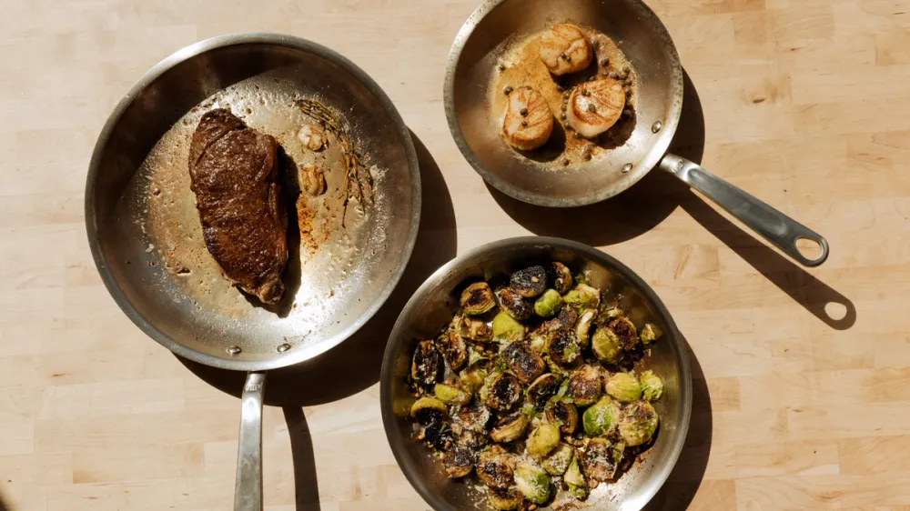 Three stainless steel pans on a wooden board holding a seared steak, seared scallops and roasted brussels sprouts, with browned residue visible in the steak pan